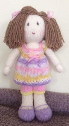 a knitted doll sitting on top of a bed next to a white wall with a pink, yellow and purple striped dress