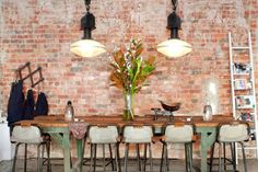 a dining room table and chairs in front of a brick wall with hanging lights above it