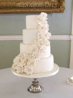 a three tiered white wedding cake with flowers on the top and bottom, sitting on a table in front of a framed sign