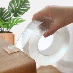 a person holding a piece of clear tape over a wooden table with a plant in the background