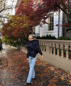 an older woman walking down the sidewalk in front of some trees with leaves on it