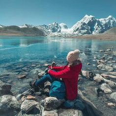 a person sitting on some rocks by the water with mountains in the backgroud