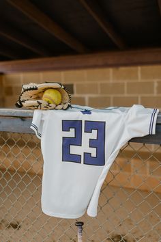 a baseball glove and ball sitting on top of a white jersey hanging from a fence