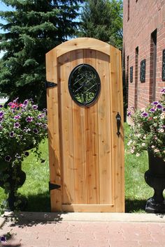 a wooden door with a clock on it in front of some plants and flowers near a brick building