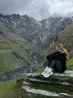 a woman sitting on top of a wooden bench next to a lush green hillside covered in snow capped mountains