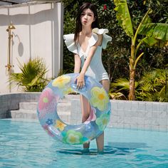 a woman holding an inflatable flowered pool float near a swimming pool with trees and bushes behind her