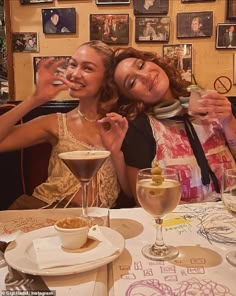 two women sitting at a table with drinks and pictures on the wall in the background