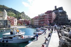 people are walking on the dock near boats in the water and buildings along the shore