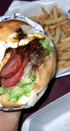 a hand holding a sandwich with meat, cheese and lettuce next to french fries