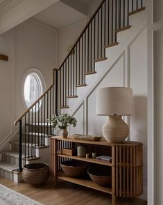 a wooden table sitting under a stair case next to a lamp and vase on top of a hard wood floor