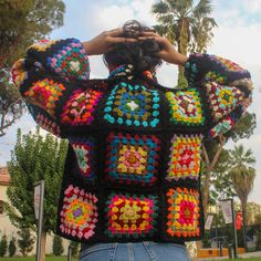 a woman wearing a colorful crochet sweater with her hands on her head, standing in front of palm trees