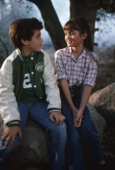 two young boys sitting on top of a rock in front of some trees and rocks