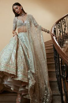 a woman in a white and gold lehenga is standing on the stairs with her hands behind her back