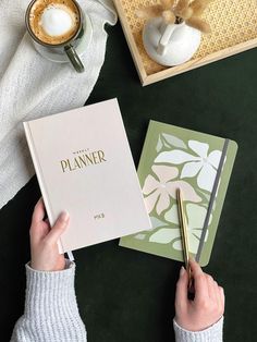 a person holding a pen and writing on a planner next to a cup of coffee