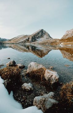 the water is clear and there are snow on the rocks in the foreground with mountains in the background