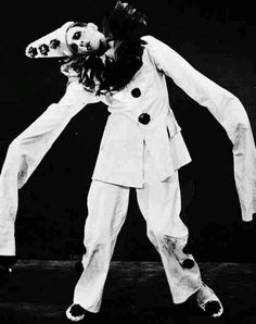 a man in white suit and clown makeup is holding his head with both hands while standing on one leg