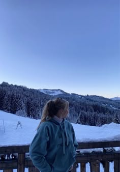 a woman standing on top of a snow covered slope next to a wooden fence with trees in the background