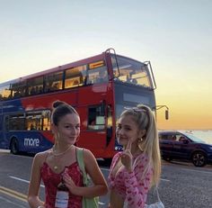 two girls standing in front of a double - decker bus on the side of the road