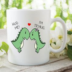 two green dinosaurs touching noses on a white coffee mug with the words hug me, i'm trying