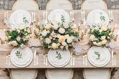 the table is set with white plates and silverware, pink flowers and greenery