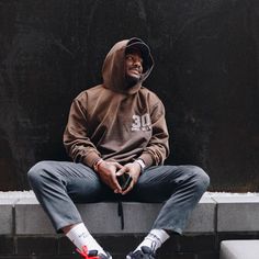 a man in a brown hoodie sitting on a cement bench with his feet crossed