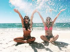 two women in bikinis are sitting on the beach and splashing water from their hands