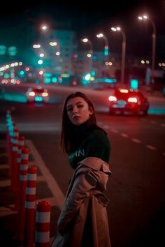 a woman standing on the side of a road at night with traffic lights in the background