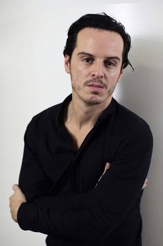 a man with his arms crossed leaning against a wall and looking at the camera while wearing a black shirt