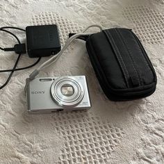 a compact camera sitting on top of a bed next to a black case and charger