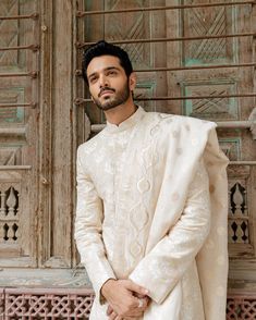 a man standing in front of a wooden door wearing a white suit and tie with an intricate pattern on it