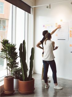 two people standing in front of a whiteboard with sticky notes on it and plants