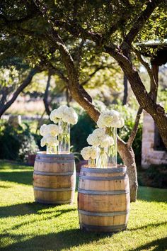 two wooden barrels with vases filled with white flowers on the grass near some trees