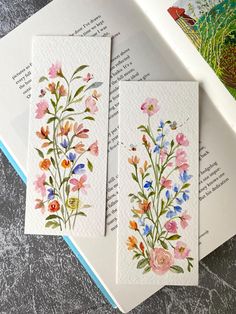 three cards with watercolor flowers on them sitting on top of a table next to an open book