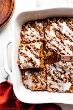 a white casserole dish filled with cinnamon rolls and topped with icing next to a wooden spoon