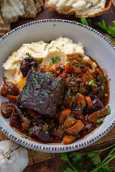 a white bowl filled with meat and potatoes on top of a wooden table next to bread