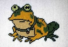 a cross stitch frog sitting on top of a white sheet with green eyes and legs