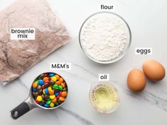 ingredients to make m & m's laid out on a white counter top, including eggs, flour, milk, and chocolate