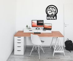 a desk with two computers on it in front of a white wall and a black backpack