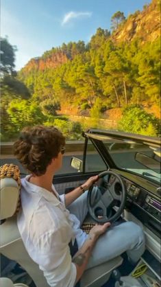 a woman driving a car down a road next to a lush green forest covered hillside