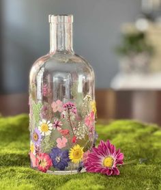 a glass bottle with flowers painted on it sitting on a moss covered tablecloth next to a pink flower