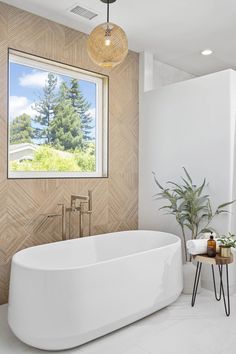 a white bath tub sitting under a window next to a wooden table and potted plant