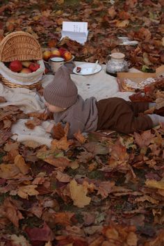 a small child laying on the ground with leaves around him and apples in a basket