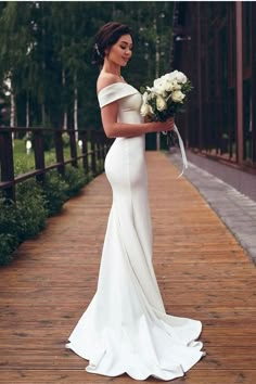 a woman in a white wedding dress holding flowers on a wooden walkway near some trees