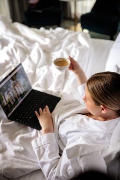 a woman is laying in bed with her laptop and cup of coffee while she looks at the screen