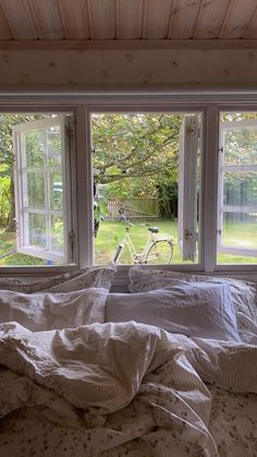 an unmade bed in front of three windows with bikes out the window behind it