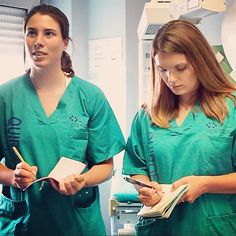 two women in green scrubs are writing on paper and looking at something while standing next to each other