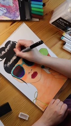 a person is drawing on a piece of paper at a table with markers and pens