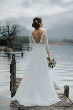 a woman in a wedding dress standing on a dock looking out at the water with her back to the camera