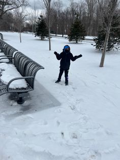 a young child standing in the snow next to a park bench with their arms outstretched