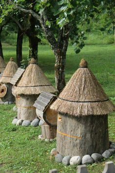 small huts made out of logs and grass in the park with trees on either side